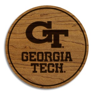 May include: A round wooden coaster with a dark brown border and a lighter wood grain center. The center features the letters "GT" stacked, with "GEORGIA TECH" below. The coaster is a decorative item.
