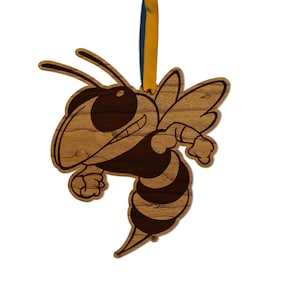 May include: Wooden ornament in the shape of a cartoon hornet, with a smiling face and striped body. The ornament is made of light brown wood with dark brown details and a yellow and blue ribbon for hanging.