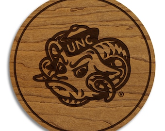 May include: A round wooden coaster with a dark brown engraved design. The design features a ram's head with the letters "UNC" on its hat. The coaster has a natural wood grain pattern and a dark brown border.