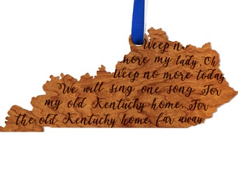 May include: A wooden ornament shaped like the state of Kentucky with the lyrics to the song "My Old Kentucky Home" engraved on it. The ornament is brown and has a blue ribbon attached to the top.