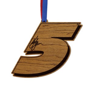 May include: Wooden ornament in the shape of the number 5, with a dark outline and a wood grain texture. The ornament has a signature on the front and is suspended by a blue and red ribbon.