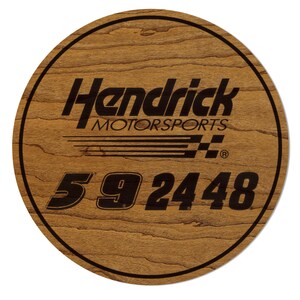 May include: A round wooden sign with the text "Hendrick Motorsports" in black, along with the numbers "59 24 48". The sign has a dark brown border and a wood grain background.