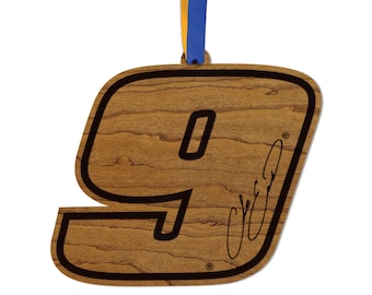 May include: Wooden ornament in the shape of the number 9, with a wood grain finish and a black outline. The number 9 has a black outline and a signature. A blue and yellow ribbon is attached for hanging.