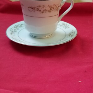May include: A white teacup and saucer with a delicate floral pattern in brown and gold. The cup has a handle and the saucer has a scalloped edge.