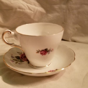 May include: A white teacup and saucer set with a floral design featuring pink roses and green leaves. The set has a gold trim around the edges.