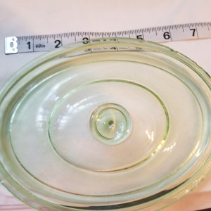Könnte beinhalten: Ein durchsichtiger Glasdeckel mit grünem Farbton und einem erhabenen Knopf in der Mitte. Der Deckel ist oval geformt und hat eine glatte, polierte Oberfläche.