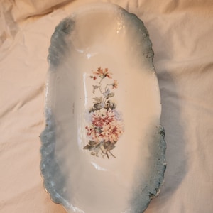 Op de afbeelding: Ovale, antieke keramische schaal met een bloemenontwerp. De schaal heeft een wit interieur met een geschilderd boeket van roze en rode bloemen. De rand heeft een blauwgroene, getextureerde rand, wat een vintage esthetiek aan het stuk toevoegt.