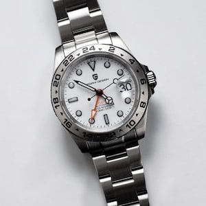 Può includere: Orologio automatico Pagani Design GMT in acciaio inossidabile con quadrante bianco. L'orologio presenta una lunetta nera con numeri bianchi, un datario a ore 3 e una lancetta GMT arancione. Resistente all'acqua fino a 100 metri.