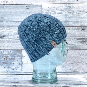 Puede incluir: Un gorro azul de punto con un patrón de punto de cable y una etiqueta de cuero marrón que dice "TKG".