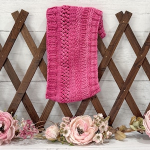 May include: A vibrant pink knitted scarf with a textured design. The scarf features a central cable knit pattern and ribbed edges. The scarf is displayed against a wooden trellis background, with floral accents at the base.