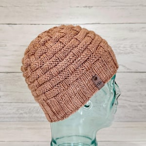 Puede incluir: Un gorro de punto color marrón claro con un patrón de tejido de cesta texturizado en la corona y un borde acanalado. Una pequeña etiqueta rectangular de cuero marrón con las letras TKG en relieve está unida al lado del borde acanalado.
