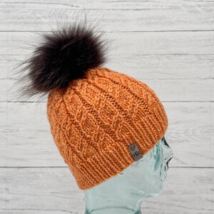 May include: An orange knitted hat with a diamond pattern and a dark brown fur pom-pom. The hat has a small tag with the letters "TKG". The hat is made of a soft, warm material, perfect for cold weather.
