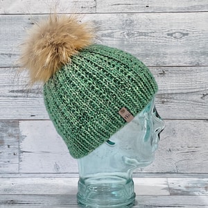 Könnte beinhalten: Eine grüne Strickmütze mit einem Kunstfellbommel. Die Mütze hat ein braunes Leder-Etikett mit den Buchstaben "TKG" darauf.