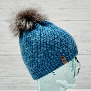May include: A blue knitted beanie with a fluffy brown pom pom on top. The beanie has a small brown leather tag with the letters "TKG" on it.