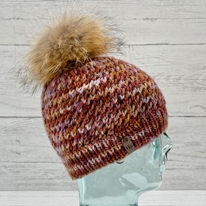 May include: A brown, pink, and orange knit beanie with a fluffy brown pom pom. The beanie has a brown leather tag with the text "TKG" on it.
