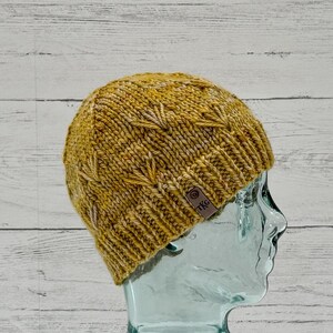 May include: Mustard yellow knit beanie with a textured leaf pattern. The hat has a folded brim and a small leather tag with the letters "TKG". The hat is displayed on a clear glass head form.