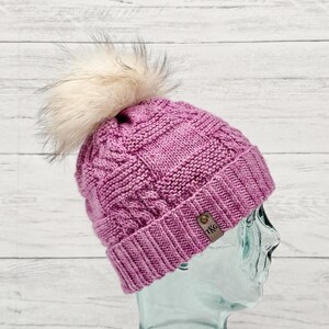 Può includere: Un berretto magenta lavorato a maglia con un motivo a trecce e un soffice pompon color crema. Il cappello ha una piccola etichetta in pelle marrone con le iniziali "TKG".