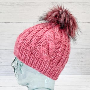 May include: A pink knitted beanie with a cable knit pattern and a black and pink faux fur pom pom.