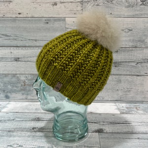 May include: A knitted olive green beanie with a ribbed design and a large, fluffy, light gray pom-pom. The hat has a small, brown leather tag with the initials "TKG". The hat is displayed on a glass head.
