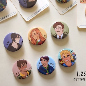 May include: A collection of 1.25" button pins featuring colorful illustrations of various characters. Each pin showcases a different character design with unique facial features and clothing. The pins are arranged on a light-colored surface.