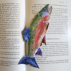 May include: A bookmark shaped like a rainbow trout with a vibrant green, pink, and blue speckled pattern. The fish is depicted with its mouth open, featuring detailed scales and fins, resting against the open pages of a printed book.