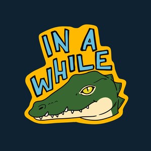 May include: A graphic design featuring a green alligator head with a yellow eye and white teeth. The alligator is set against a yellow background with the words "IN A WHILE" in blue, all on a navy blue background.