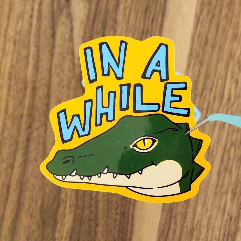 May include: A yellow sticker with the words "IN A WHILE" in blue block letters. The sticker features a green alligator head with a yellow eye and white teeth. The sticker is being held by a pair of blue tweezers.