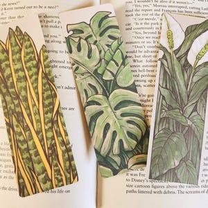 May include: Three botanical-themed bookmarks rest on open book pages. The bookmarks feature illustrations of a snake plant, monstera leaves, and a peace lily. The illustrations are in shades of green and white.