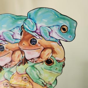 K&ouml;nnte beinhalten: Ein gestanzter Aufkleber mit einem vertikalen Stapel aus f&uuml;nf illustrierten Fr&ouml;schen. Der oberste Frosch ist hellblau, der mittlere hellbraun und der unterste gr&uuml;n, wobei weitere Froschk&ouml;pfe im Stapel zu sehen sind. Jeder Frosch hat dunkle Augen.