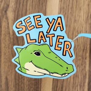 May include: A sticker with a cartoon alligator head and the words "SEE YA LATER" in orange letters on a light blue background. The alligator is green with a white belly and a smiling expression.
