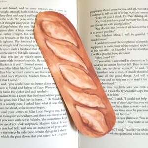 Puede incluir: Un marcapáginas con forma de barra de pan, que presenta una ilustración realista de un pan dorado con marcas de corte diagonales. El marcapáginas está colocado verticalmente sobre las páginas abiertas de un libro con texto negro.