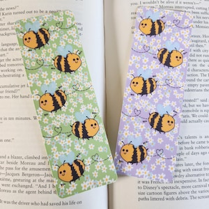 Könnte beinhalten: Zwei rechteckige Lesezeichen mit einem sich wiederholenden Muster von Cartoon-Bienen. Eines hat einen grünen Hintergrund mit weißen Blumen, das andere einen lila Hintergrund mit weißen Blumen. Die Bienen sind gelb und schwarz.