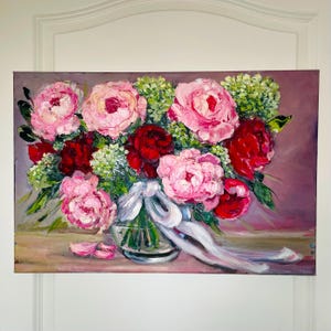 Pode incluir: Uma pintura a óleo sobre tela representando um buquê de peônias rosas e vermelho-escuras com folhagem verde em um vaso de vidro transparente. Uma fita clara está amarrada ao redor do vaso e várias pétalas rosas estão espalhadas na superfície.