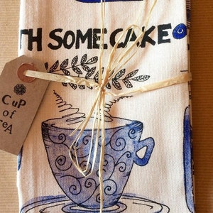 Puede incluir: Un paño de cocina blanco con ilustraciones azules y negras de tazas de té y el texto "With some cake and a cup of tea". El paño de cocina está atado con un lazo de cuerda marrón.