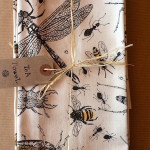 May include: A set of tea towels with a white background and black and yellow insect illustrations. The towels are tied together with twine and a brown tag that reads "Tea Towel".