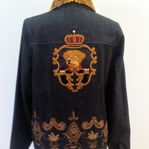 May include: A dark blue denim jacket with a gold embroidered crest featuring a dog wearing a crown. The crest is surrounded by a gold frame and the jacket has a gold embroidered border at the bottom.