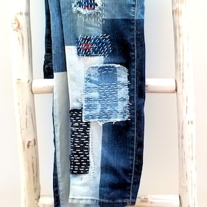 May include: A pair of blue denim jeans with multiple patches sewn on. The patches are made of different shades of blue denim and have white stitching. The jeans are hanging on a white wooden ladder.