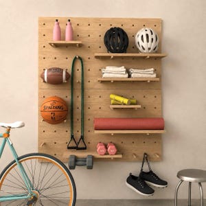 Op de afbeelding: Een lichthouten pegboard wandorganizer met planken en haringen met twee roze flessen, een zwarte helm, een witte helm, gevouwen witte handdoeken, een gele foamroller, een rode yogamat, roze dumbbells, een football, een basketbal en schoenen.