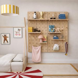May include: A light wood pegboard wall organizer with multiple adjustable shelves holding various toys, a trophy, a small lamp, and a pink cloth. The unit features a grid of circular holes for shelf placement and is mounted on a plain wall.