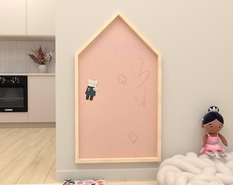 May include: A house-shaped pink chalkboard with a light wood frame is mounted on a wall. The chalkboard has drawings and a bear-shaped magnet. A doll in a pink dress sits on a white cloud-shaped cushion.