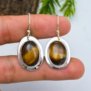 May include: A pair of oval-shaped earrings with a silver frame and a polished, golden-brown stone center. The earrings have a simple hook design for pierced ears. The stones have a natural, tiger-eye appearance.