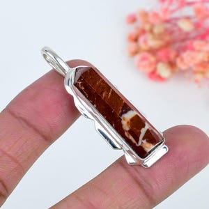Pode incluir: Um pingente com uma pedra longa de corte hexagonal em tons de castanho e branco, engastada numa armação de metal prateado polido. A pedra tem um acabamento liso e brilhante com variações de cor distintas e um orifício no topo.