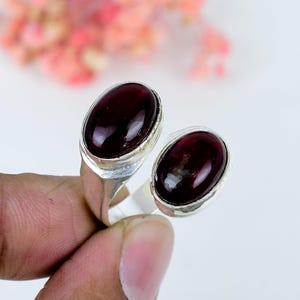 Pode incluir: Um anel de prata tipo bypass com duas pedras preciosas granada vermelho-escuras de formato oval, fixadas em engastes de metal liso. O anel tem um design aberto com uma banda de prata polida que se curva ao redor do dedo, exibindo as pedras.