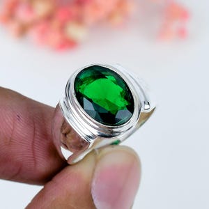 Könnte beinhalten: Ein Silberring mit einem großen, oval geschliffenen, leuchtend grünen Edelstein in einer polierten Metallschiene. Der facettierte Stein ist sicher in einer glatten, abgerundeten Zargenfassung gefasst, die die tiefe smaragdgrüne Farbe betont.