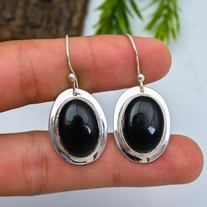 May include: Silver drop earrings featuring oval, polished black stones. Each stone is set in a silver frame and hangs from a simple hook. The earrings are shown against a neutral backdrop.