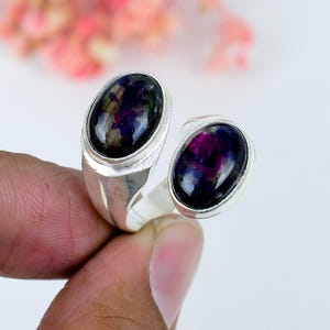 Puede incluir: Un anillo de plata de diseño abierto con dos cabujones ovalados de superficie oscura e iridiscente, que muestran tonos profundos de morado, azul y negro. La banda de metal pulido está abierta y sujeta las piedras en engastes redondeados.