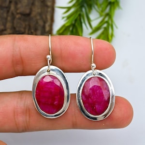 Op de afbeelding: Een paar zilveren oorbellen met ovale, dieprode edelstenen. De edelstenen zijn gezet in een zilveren bezel en de oorbellen hebben een eenvoudig haakontwerp. De oorbellen worden gepresenteerd tegen een neutrale achtergrond.