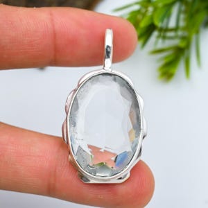 May include: A clear, oval gemstone pendant in a silver setting. The gemstone has internal facets that reflect light, creating a rainbow effect. The pendant is held by a person's fingers.
