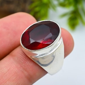 May include: A silver ring featuring a large, oval-cut, deep red gemstone. The gemstone is set in a polished silver band. The ring is shown close-up, highlighting the facets of the stone and the smooth metal.
