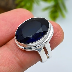 Peut inclure: Une bague en argent ornée d'une grande pierre précieuse ovale bleu foncé. La pierre est taillée à facettes, captant la lumière et affichant un éclat brillant. La bague a un design simple et élégant.